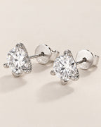 The Elio Moissanite Stud Earrings — clear round faceted studs in silver-colored prong settings with straight posts and circular S925-marked backs on pale greige, reference-based AI visualization