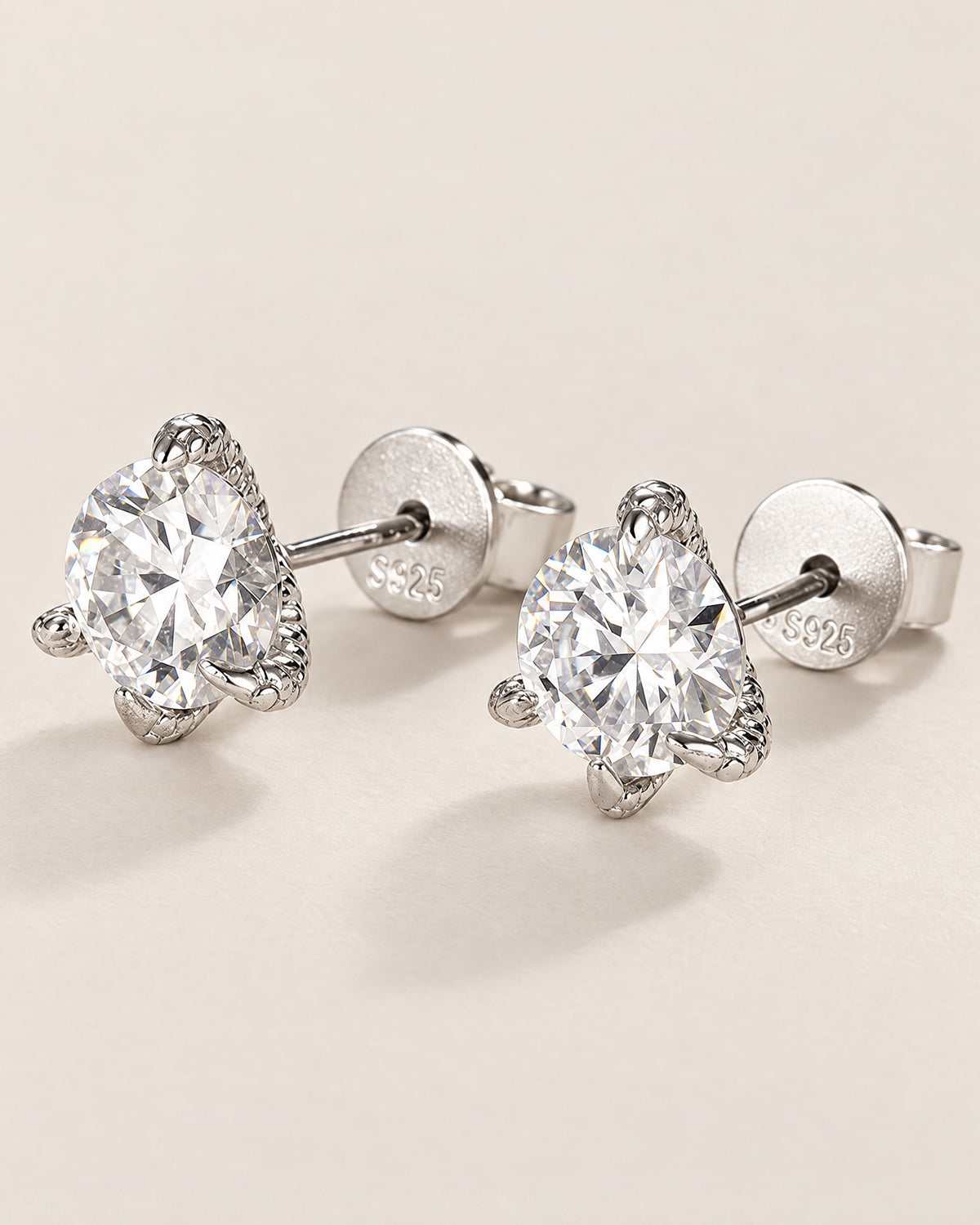 The Elio Moissanite Stud Earrings — clear round faceted studs in silver-colored prong settings with straight posts and circular S925-marked backs on pale greige, reference-based AI visualization