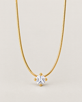 Gold-color segmented necklace in a centered open U with one clear round stone held by four rounded prongs on a pale warm greige studio surface; reference-based AI visualization.