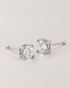 The Neva CZ Stud Earrings — silver-tone low-angle resting pair with clear round stones and visible open settings in a pale-greige studio; reference-based AI visualization.