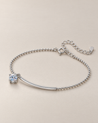 The Eden Moissanite Bracelet in polished silver tone, resting in a shallow curve with a curved bar, one round clear stone, ball-bead chain, clasp, and extension on a pale-greige background, reference-based AI visualization.