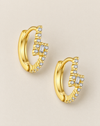 The Veyra Geometric Huggies, separated warm yellow-gold-coloured geometric huggies in a diagonal overhead flat lay with clear rectangular and round stones on pale greige, reference-based AI visualization