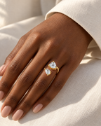 The Nessa Heart & Pear Ring in polished yellow-gold-tone metal with clear faceted heart and pear stones, correctly worn on a deep-brown adult hand against ivory linen — reference-based AI visualization.