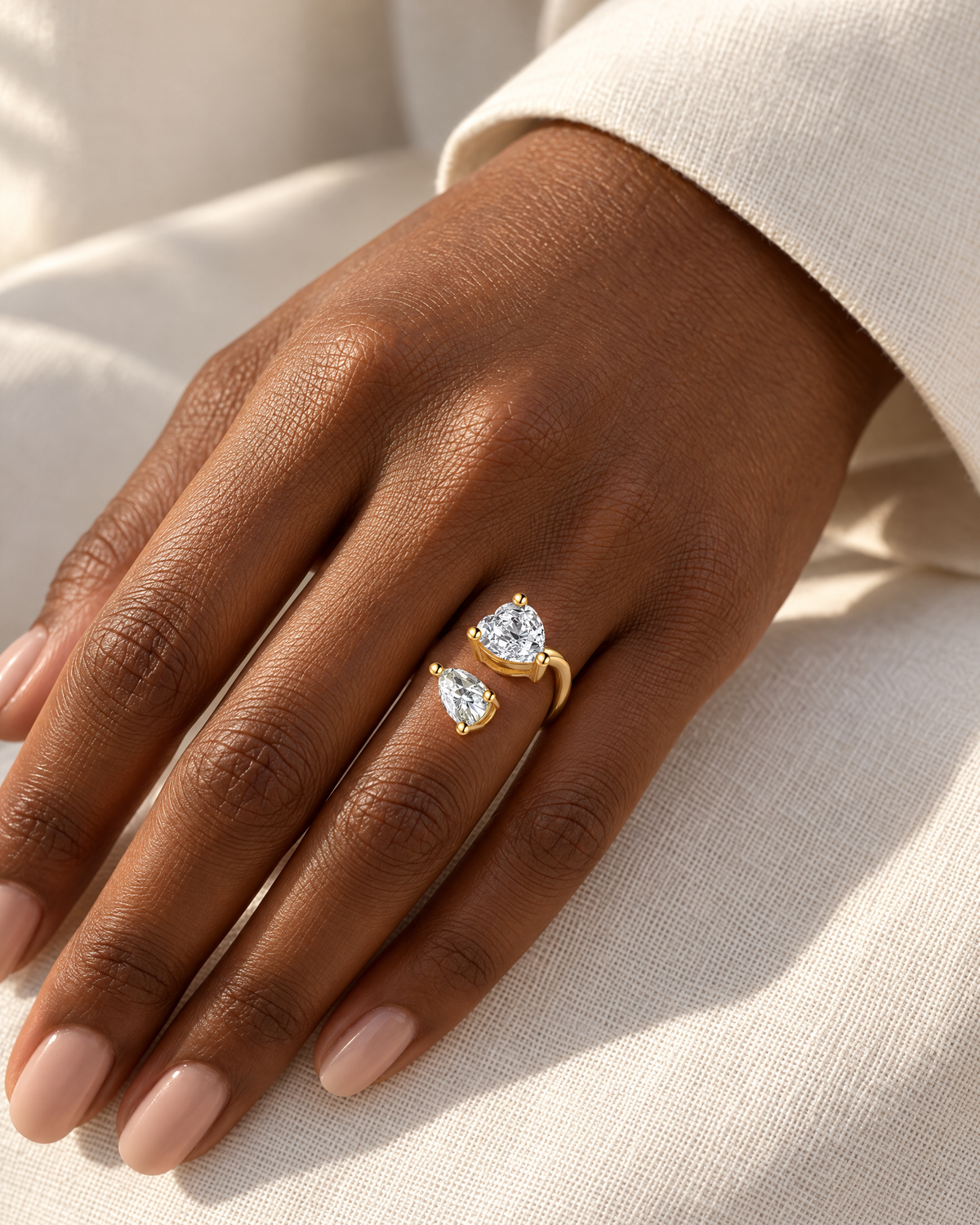 The Nessa Heart & Pear Ring in polished yellow-gold-tone metal with clear faceted heart and pear stones, correctly worn on a deep-brown adult hand against ivory linen — reference-based AI visualization.