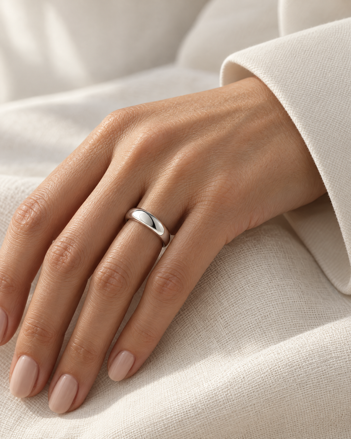 The Norden Plain Band Ring in polished silver, worn on a mature adult woman's hand with ivory linen in warm window light, reference-based AI visualization.