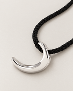 Polished crescent pendant resting on pale greige with black braided cord sweeping diagonally, reference-based AI visualization