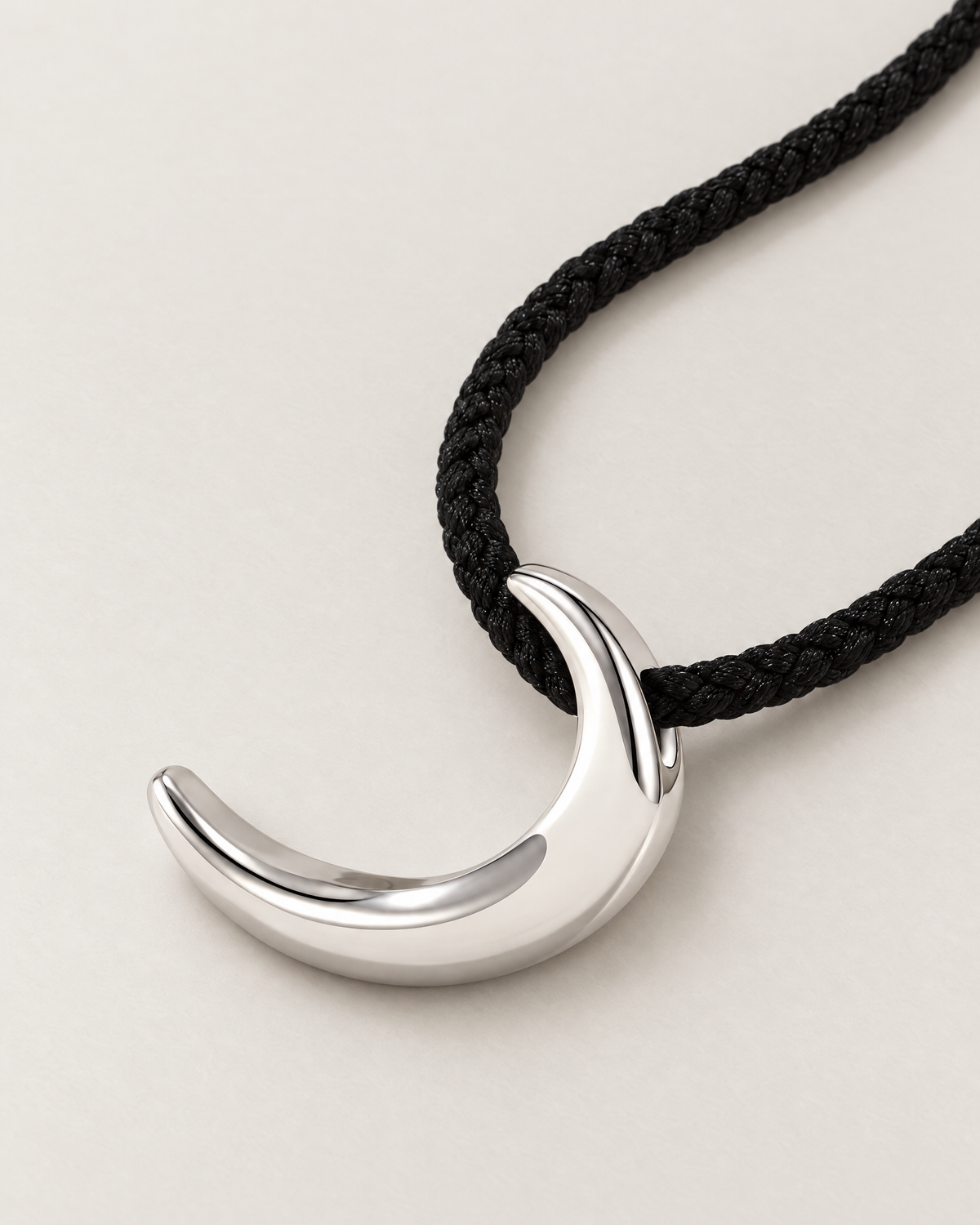 Polished crescent pendant resting on pale greige with black braided cord sweeping diagonally, reference-based AI visualization