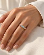 The Miri Color-Accent Band with a polished yellow-gold-colored band, three light-blue round stones and two magenta round accent stones, properly worn on a mature adult woman's finger in warm window light with ivory linen, reference-based AI visualization