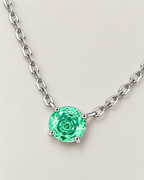 Round green faceted pendant in a silver-colored four-prong setting with linked chain arranged on a diagonal pale greige studio ground — reference-based AI visualization