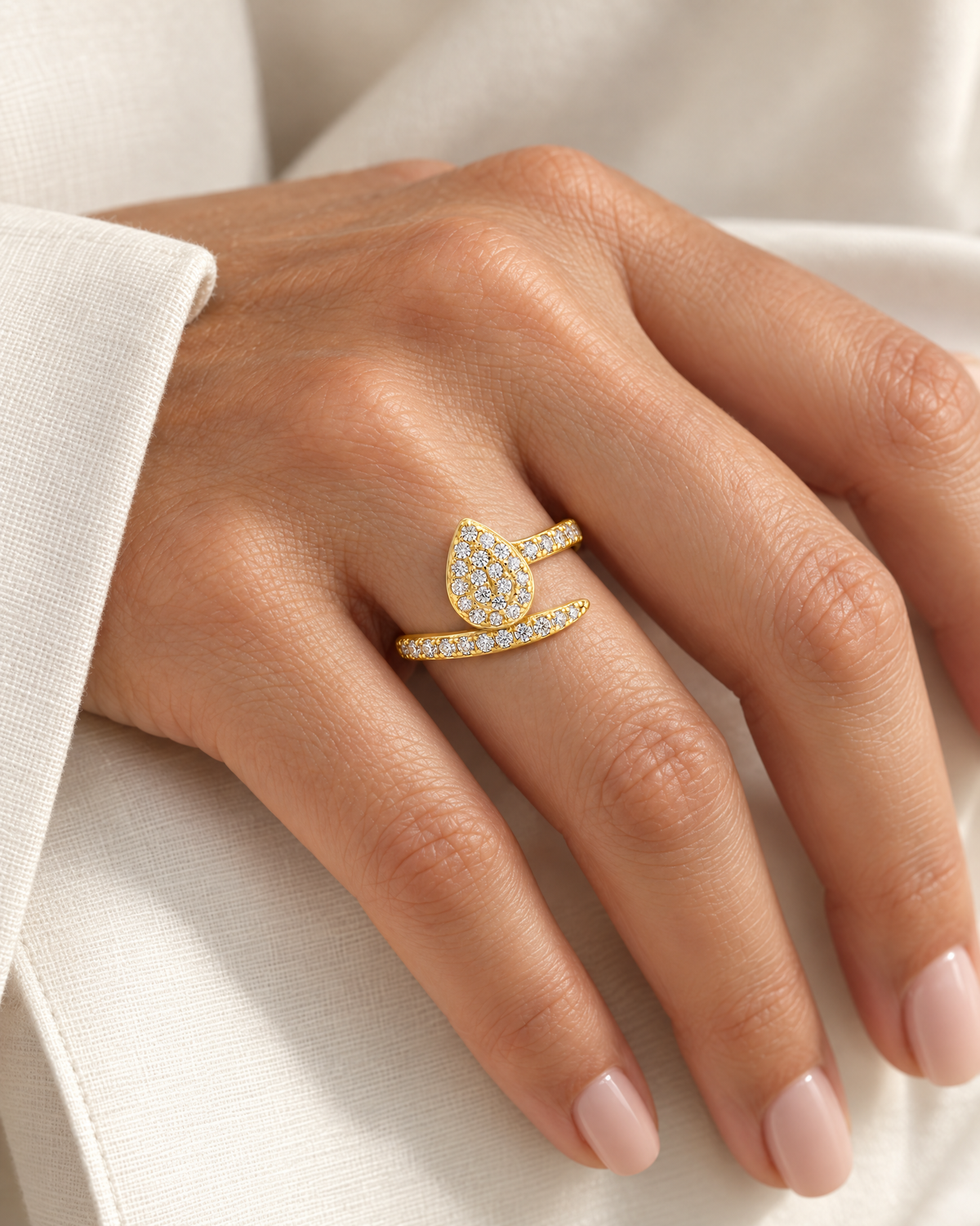 The Oona Pavé Teardrop Ring in warm polished gold with clear round pavé stones, worn on a light-warm-toned adult hand beside ivory linen; reference-based AI visualization.
