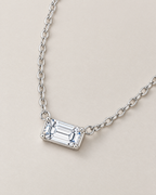 The Mara Geometric CZ Pendant — jewelry-only shallow-oblique view of the horizontal clear rectangular stone and alternating-link chain on pale warm greige; reference-based AI visualization