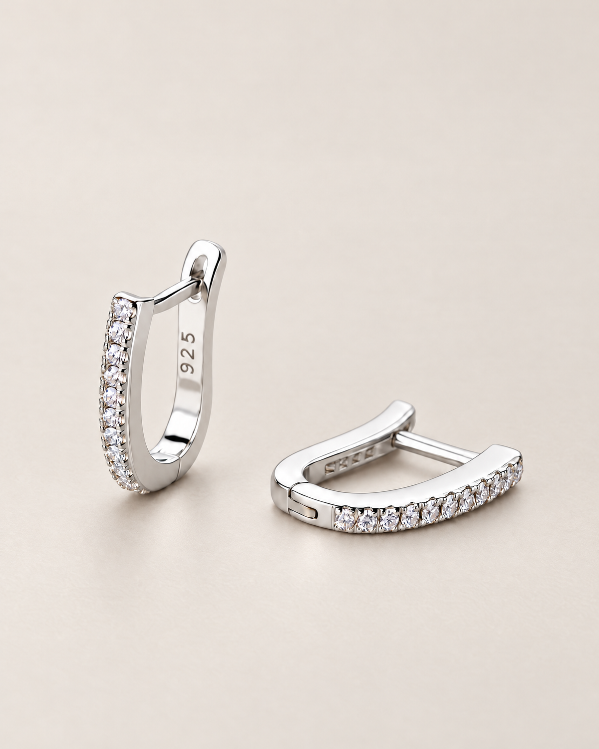 The Aria CZ Hoops — pair of polished silver-colored U-shaped hoops with clear round stone rows, one upright and one resting on pale greige, reference-based AI visualization