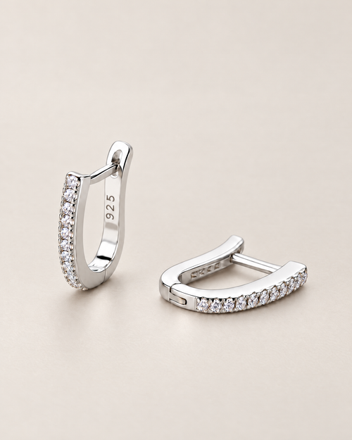 The Aria CZ Hoops — pair of polished silver-colored U-shaped hoops with clear round stone rows, one upright and one resting on pale greige, reference-based AI visualization