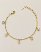 The Helle Moissanite Station Bracelet in gold tone, shown as a full five-station relaxed open curve on pale greige, reference-based AI visualization.