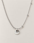 Front product-only shallow-U view of the silver-colored asymmetric necklace with a round LUCKY medallion, bead-and-bar chain at left and smooth station-bead chain at right, reference-based AI visualization