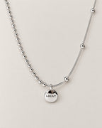 Front product-only shallow-U view of the silver-colored asymmetric necklace with a round LUCKY medallion, bead-and-bar chain at left and smooth station-bead chain at right, reference-based AI visualization