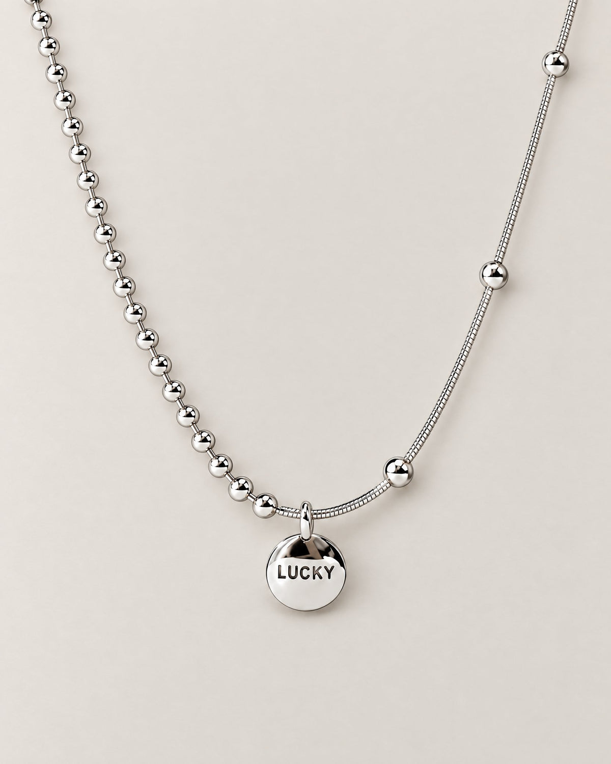 Front product-only shallow-U view of the silver-colored asymmetric necklace with a round LUCKY medallion, bead-and-bar chain at left and smooth station-bead chain at right, reference-based AI visualization