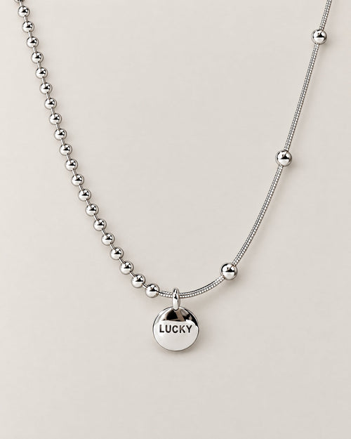 Front product-only shallow-U view of the silver-colored asymmetric necklace with a round LUCKY medallion, bead-and-bar chain at left and smooth station-bead chain at right, reference-based AI visualization