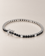 The Signe Black & White CZ Tennis Bracelet in polished silver-tone metal, shown with its open box clasp, curved safety catches, and repeating black and clear/white round stones on pale greige, reference-based AI visualization.