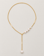 The Freya Pearl Drop Necklace — warm gold-tone cable chain in an organic oval lay with asymmetric white pearl-like accent beads and a single pearl-like drop on pale greige, reference-based AI visualization