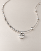 Low-oblique product-only view of a silver-colored necklace with the round LUCKY medallion and asymmetric beaded and smooth station-bead chains, reference-based AI visualization