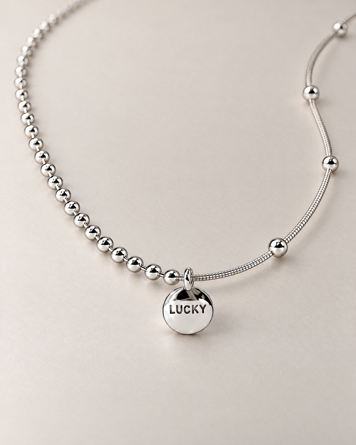 Low-oblique product-only view of a silver-colored necklace with the round LUCKY medallion and asymmetric beaded and smooth station-bead chains, reference-based AI visualization