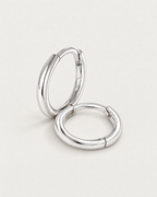 The Kiri Huggie Hoops — silver pair — leaning and resting pair on pale greige, product-only; reference-based AI visualization.