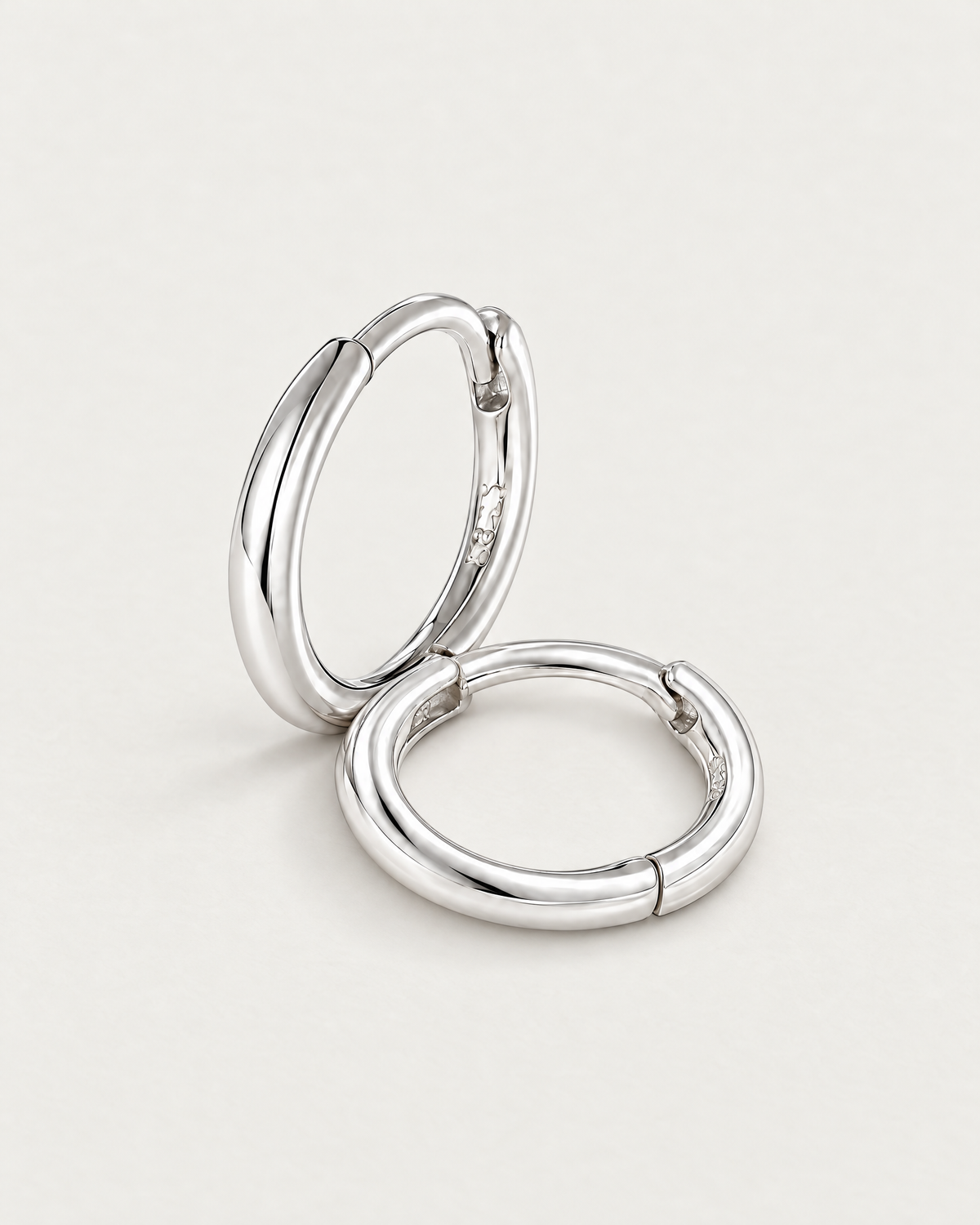 The Kiri Huggie Hoops — silver pair — leaning and resting pair on pale greige, product-only; reference-based AI visualization.