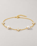 The Amelie Pearl Station Bracelet with polished yellow-gold-tone fine chain, two off-white pearl-like stations and three clear four-petal stone stations in a complete low-angle resting view on pale greige, reference-based AI visualization