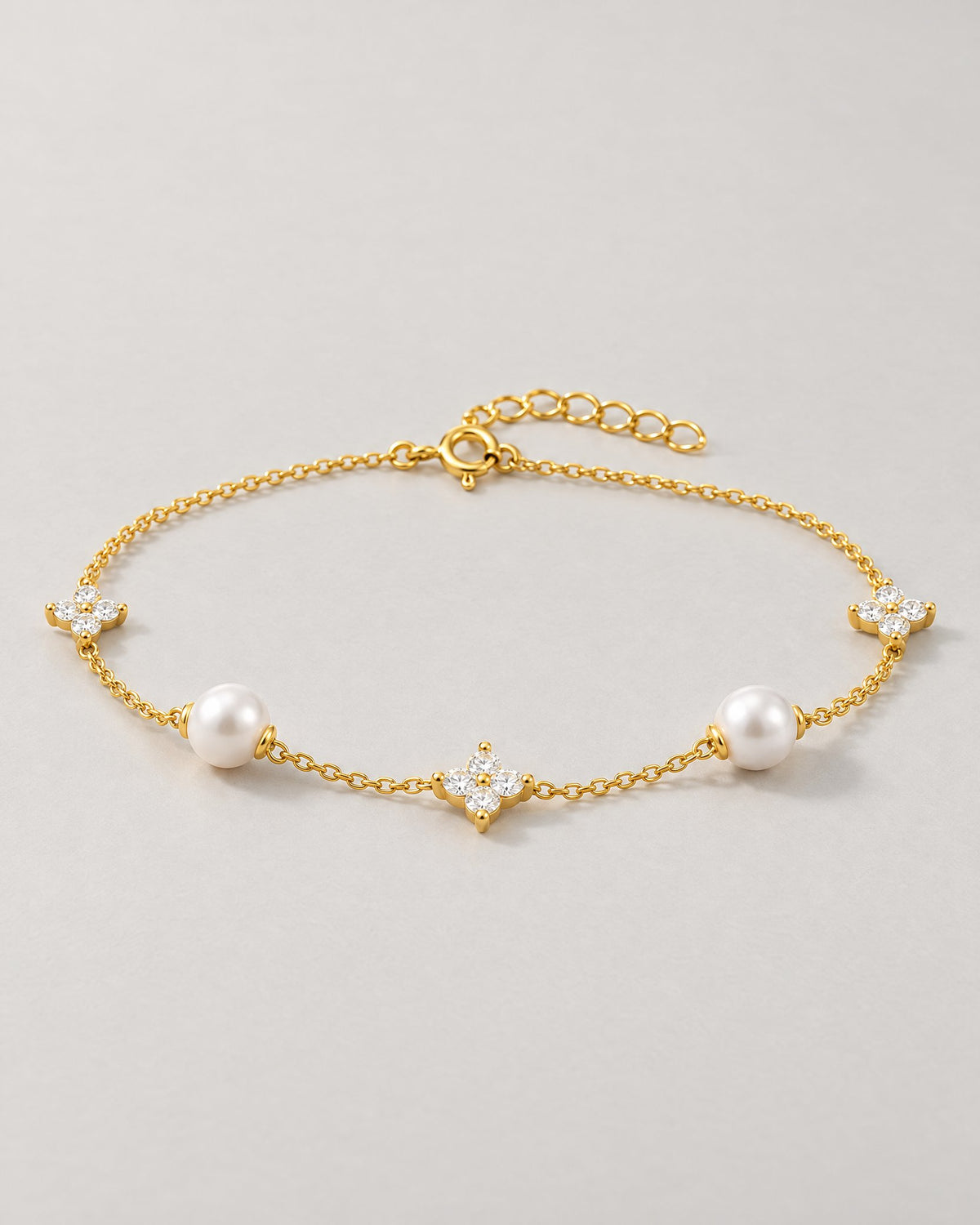 The Amelie Pearl Station Bracelet with polished yellow-gold-tone fine chain, two off-white pearl-like stations and three clear four-petal stone stations in a complete low-angle resting view on pale greige, reference-based AI visualization