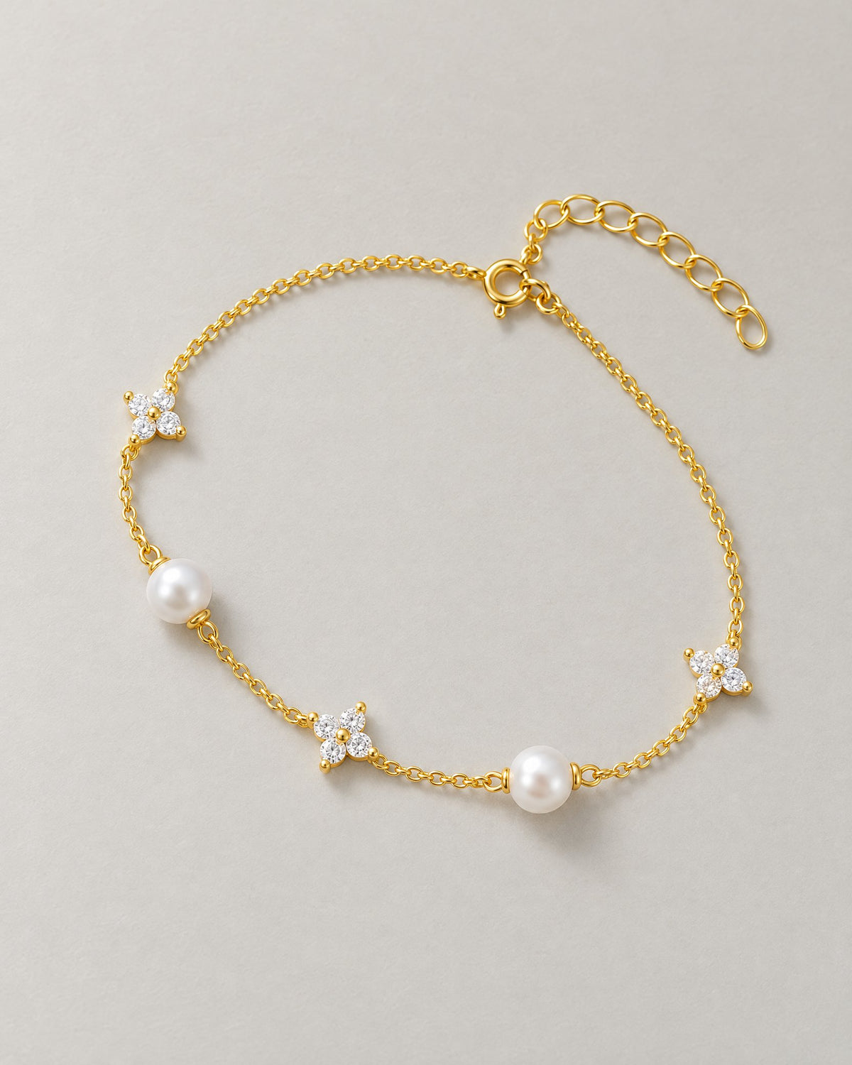 The Amelie Pearl Station Bracelet with two off-white pearl-like stations, three clear four-petal stone stations and polished yellow-gold-tone chain in a complete overhead resting lay on pale greige, reference-based AI visualization