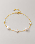 The Amelie Pearl Station Bracelet with polished yellow-gold-tone fine chain, two off-white pearl-like stations and three clear four-petal stone stations in a complete front three-quarter oval on pale greige, reference-based AI visualization