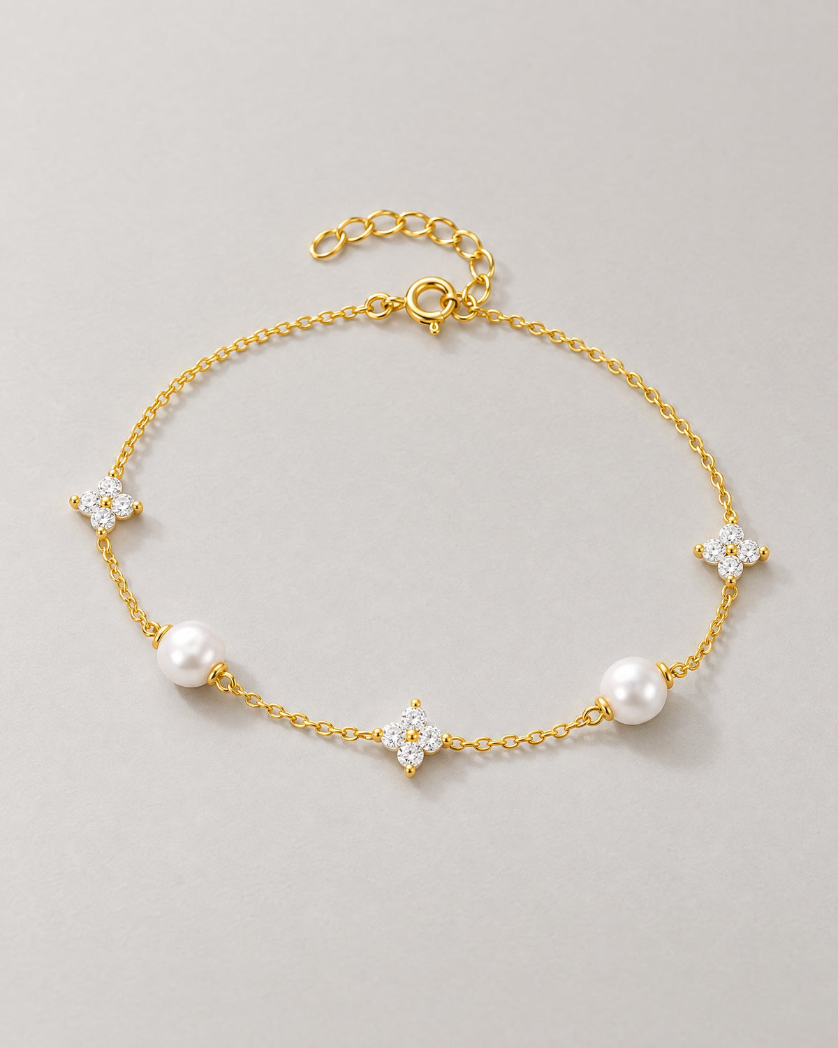 The Amelie Pearl Station Bracelet with polished yellow-gold-tone fine chain, two off-white pearl-like stations and three clear four-petal stone stations in a complete front three-quarter oval on pale greige, reference-based AI visualization