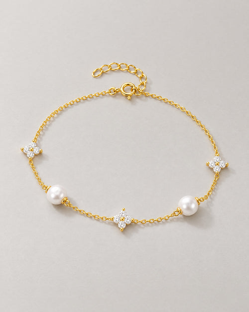 The Amelie Pearl Station Bracelet with polished yellow-gold-tone fine chain, two off-white pearl-like stations and three clear four-petal stone stations in a complete front three-quarter oval on pale greige, reference-based AI visualization