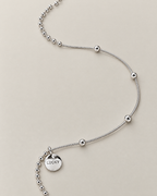 Organic flowing lay of the silver-colored necklace showing the round LUCKY medallion, close bead-and-bar chain, and smooth station-bead chain, reference-based AI visualization
