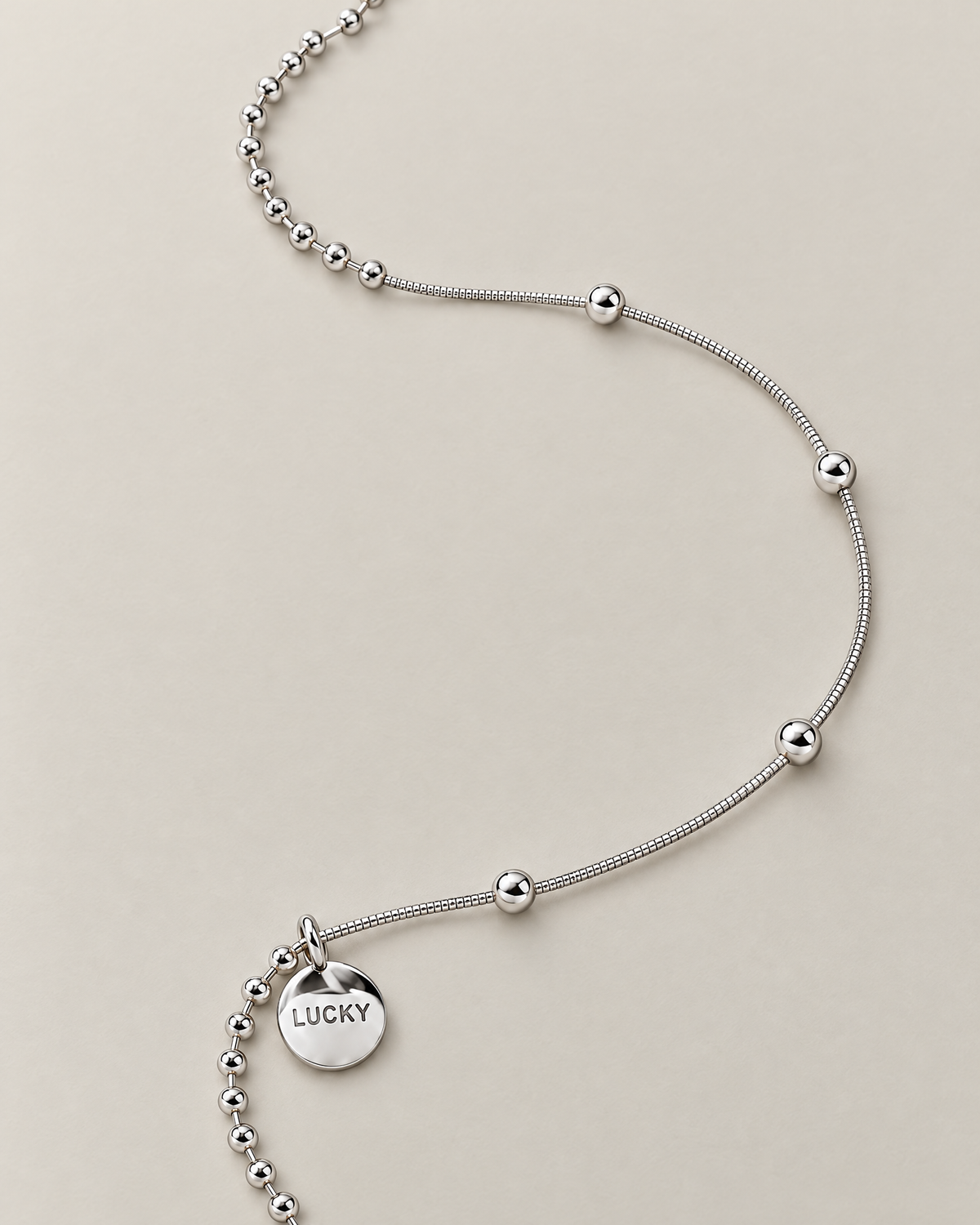 Organic flowing lay of the silver-colored necklace showing the round LUCKY medallion, close bead-and-bar chain, and smooth station-bead chain, reference-based AI visualization