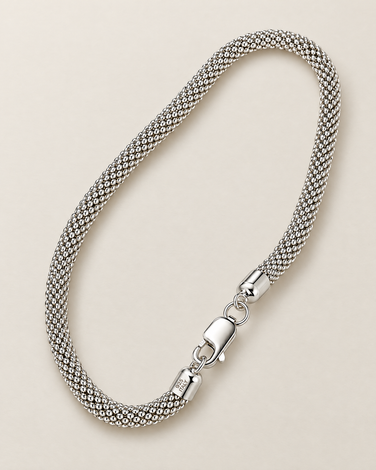 The Liva Mesh Chain Bracelet in a complete silver-tone woven-mesh diagonal oval with lobster clasp and visible 925 ITALY end-cap marking on warm pale greige; reference-based AI visualization.
