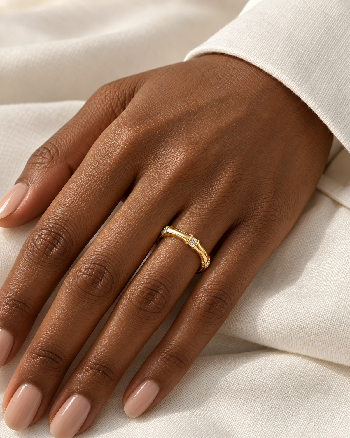 The Ebba Bamboo Band in polished yellow-gold color with clear round stone accents, worn on a deep brown adult woman's hand with ivory linen; reference-based AI visualization.