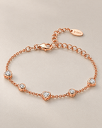 The Dahlia S925 Heart Bracelet in warm rose-gold-toned metal with five alternating clear heart and round stone stations in a low resting oval on pale greige, reference-based AI visualization.