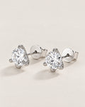 The Elio Moissanite Stud Earrings — matching clear round faceted studs in silver-colored prong settings with posts and circular S925-marked backs, front three-quarter on pale greige, reference-based AI visualization
