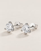 The Elio Moissanite Stud Earrings — matching clear round faceted studs in silver-colored prong settings with posts and circular S925-marked backs, front three-quarter on pale greige, reference-based AI visualization