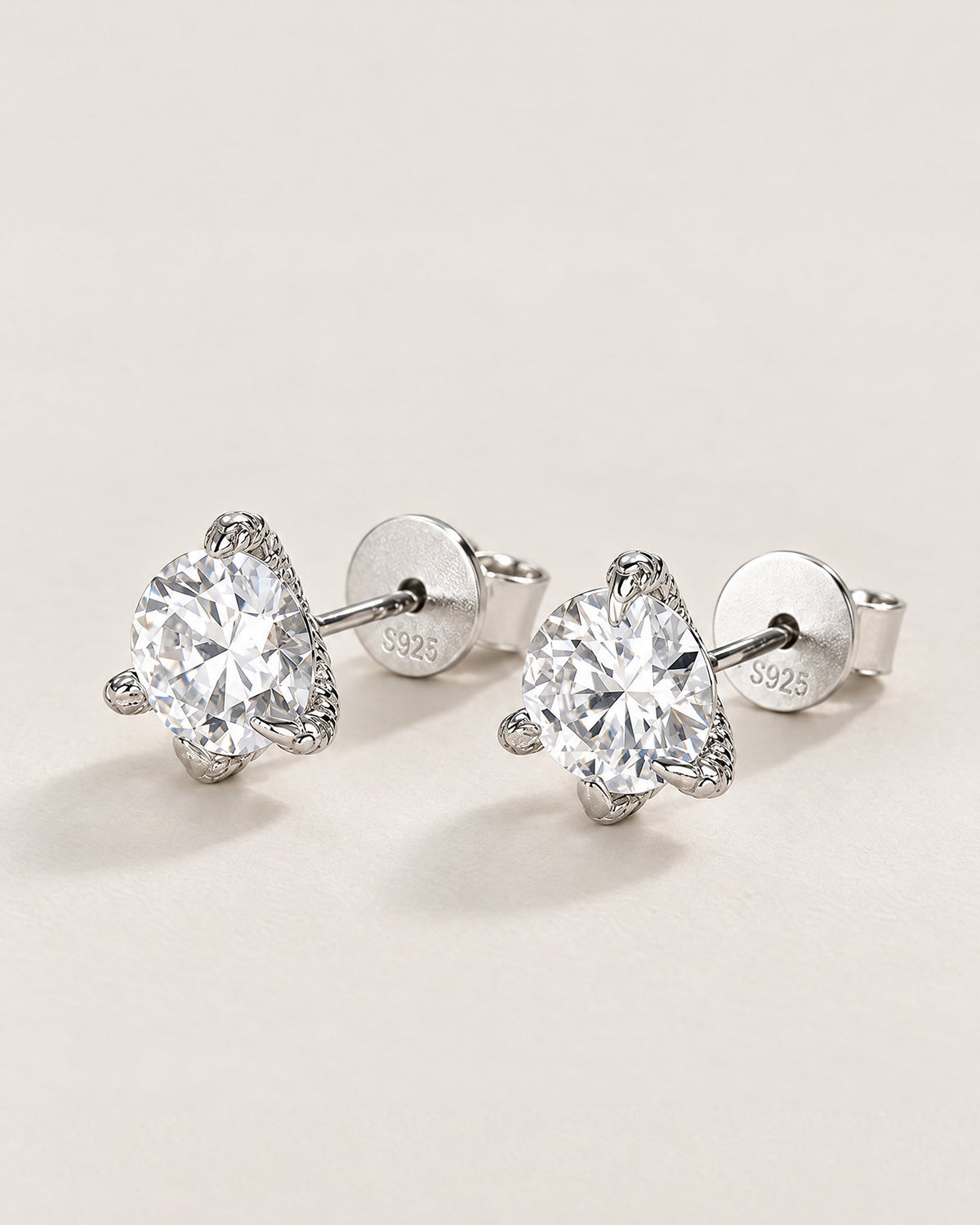 The Elio Moissanite Stud Earrings — matching clear round faceted studs in silver-colored prong settings with posts and circular S925-marked backs, front three-quarter on pale greige, reference-based AI visualization