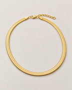 Gold-tone flat herringbone necklace in a wide rounded full loop with clasp and extender across the top on a pale greige studio ground; reference-based AI visualization
