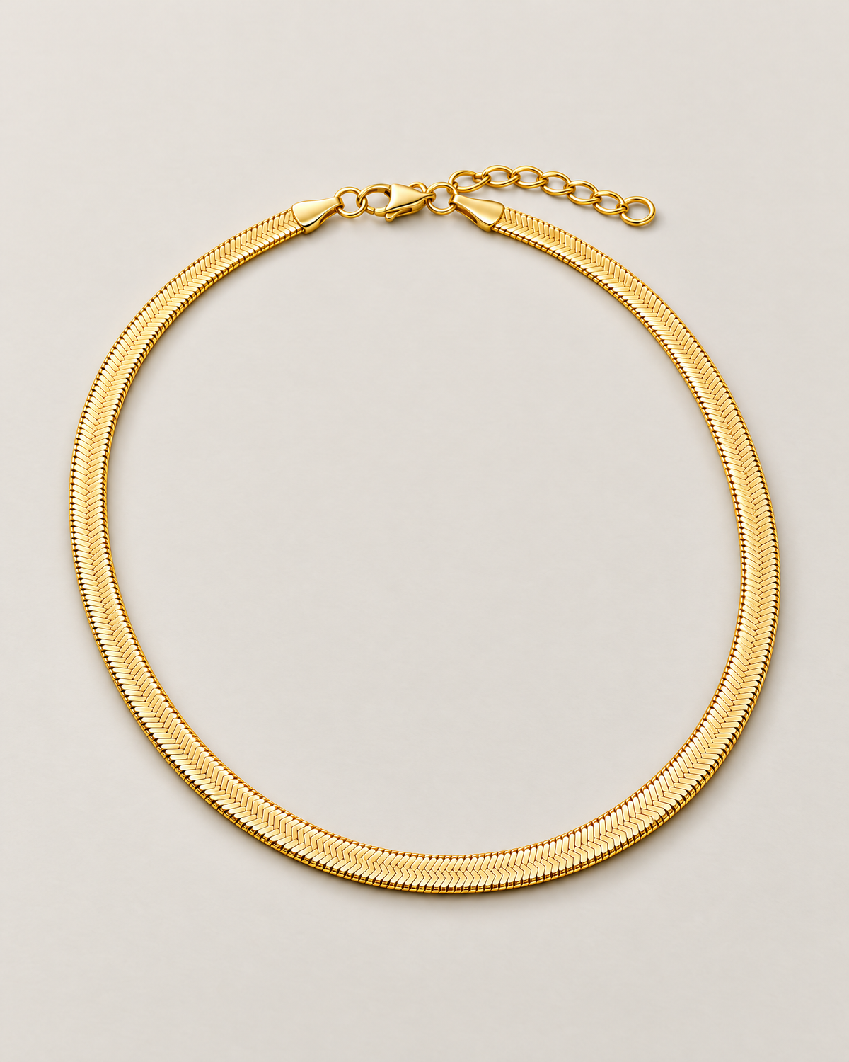 Gold-tone flat herringbone necklace in a wide rounded full loop with clasp and extender across the top on a pale greige studio ground; reference-based AI visualization