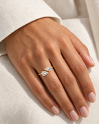 The Agnes Twin-Pear Ring in a polished yellow-gold finish with two clear pear-shaped stones, worn on a medium-warm adult hand against ivory linen — reference-based AI visualization