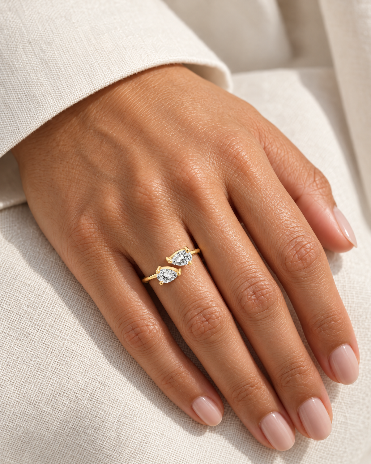 The Agnes Twin-Pear Ring in a polished yellow-gold finish with two clear pear-shaped stones, worn on a medium-warm adult hand against ivory linen — reference-based AI visualization