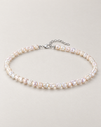 Irregular off-white pearl strand in a complete low oblique oval with visible clasp and extension on a pale greige studio surface; reference-based AI visualization