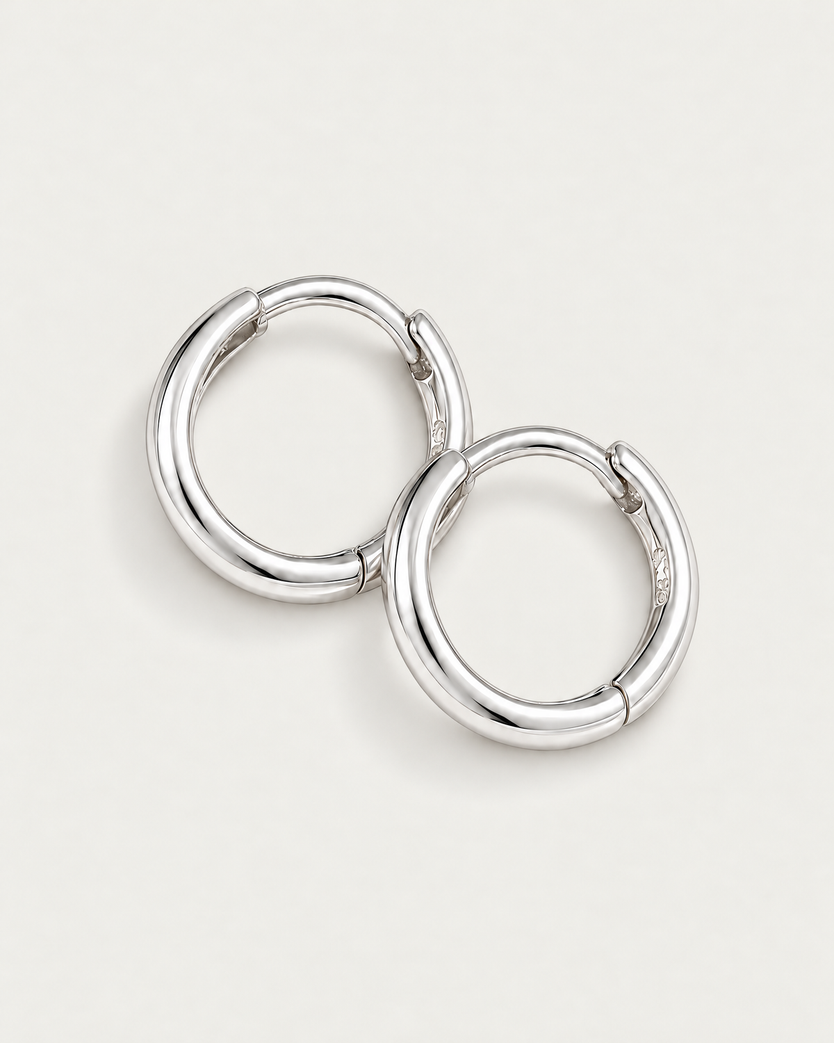 The Kiri Huggie Hoops — silver pair — diagonal flat lay on pale greige, product-only; reference-based AI visualization.