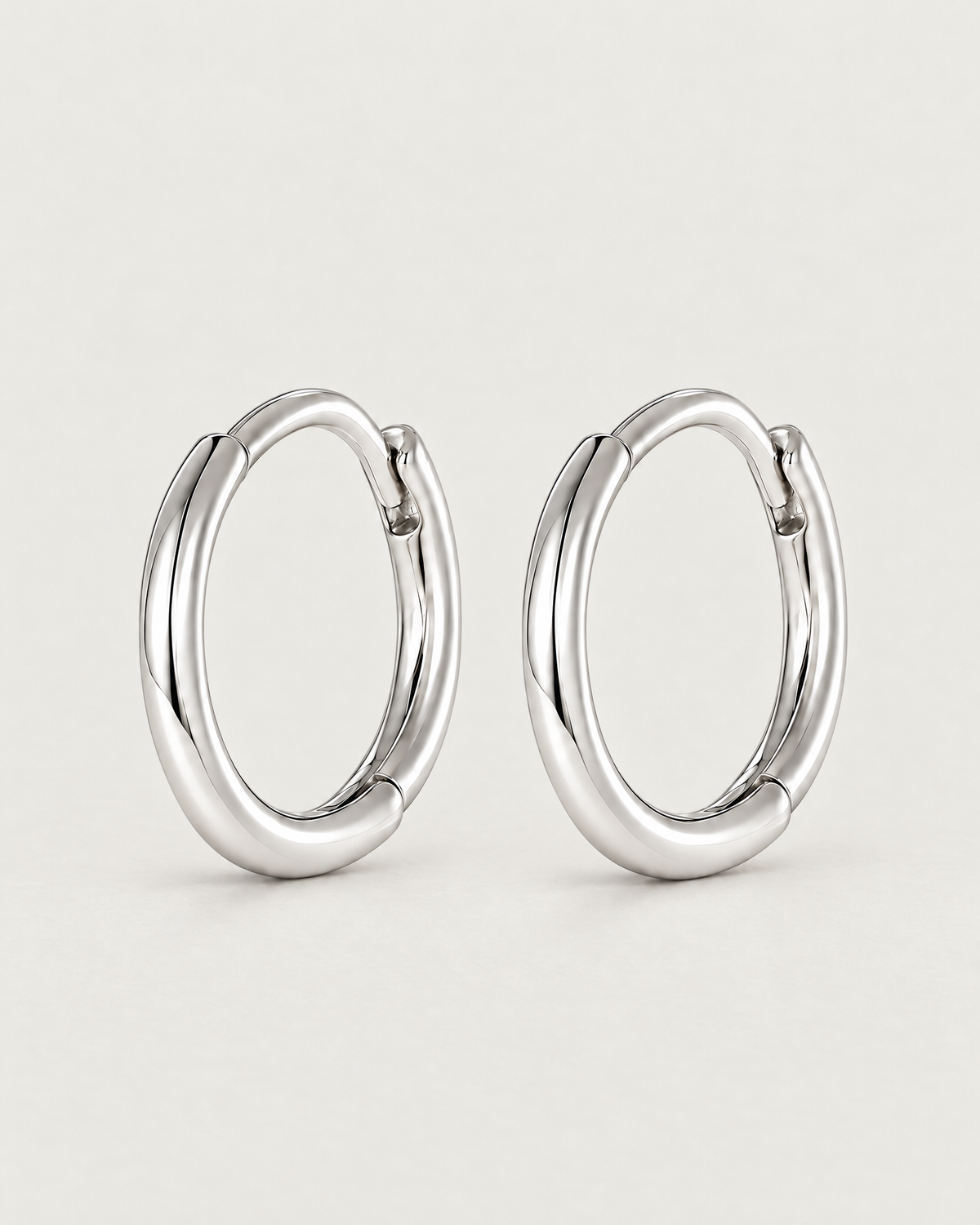 The Kiri Huggie Hoops — silver pair — angled standing pair on pale greige, product-only; reference-based AI visualization.
