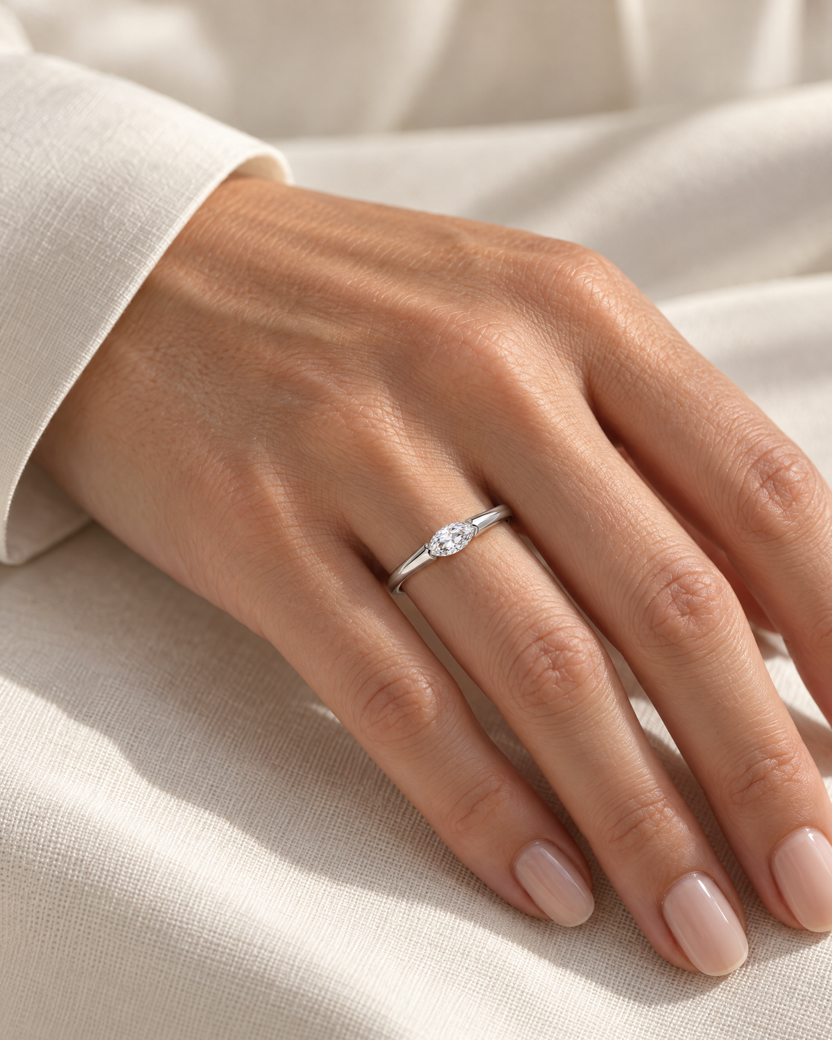 The Aure Marquise-Cut CZ Ring, polished silver marquise-cut ring worn on a light-warm adult hand with ivory linen in warm window light — reference-based AI visualization.